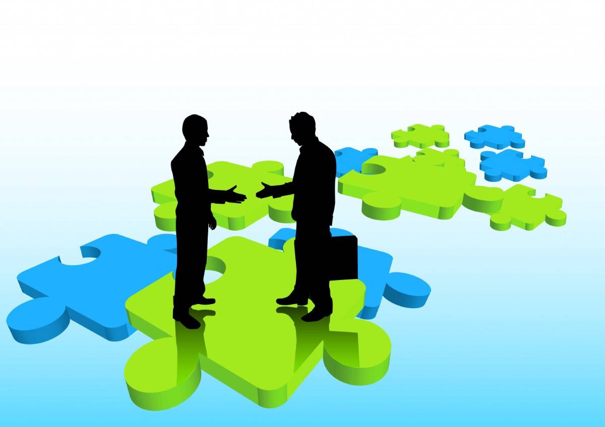 Image of 2 business people extending hands on green and blue puzzle pieces.