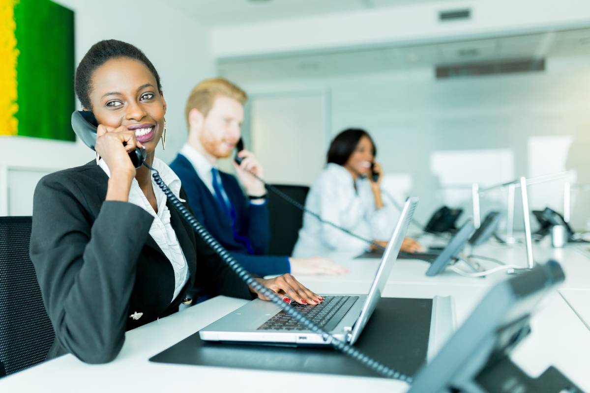 A woman using a laptop and talking on the phone, with two of her coworkers in the background