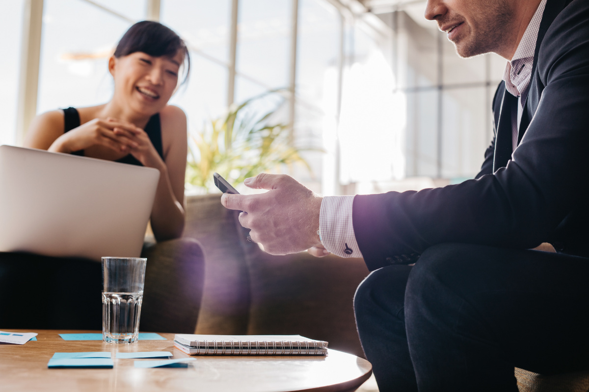 Businessman Checking Cell Phone with Smiling Woman on Latop in Background